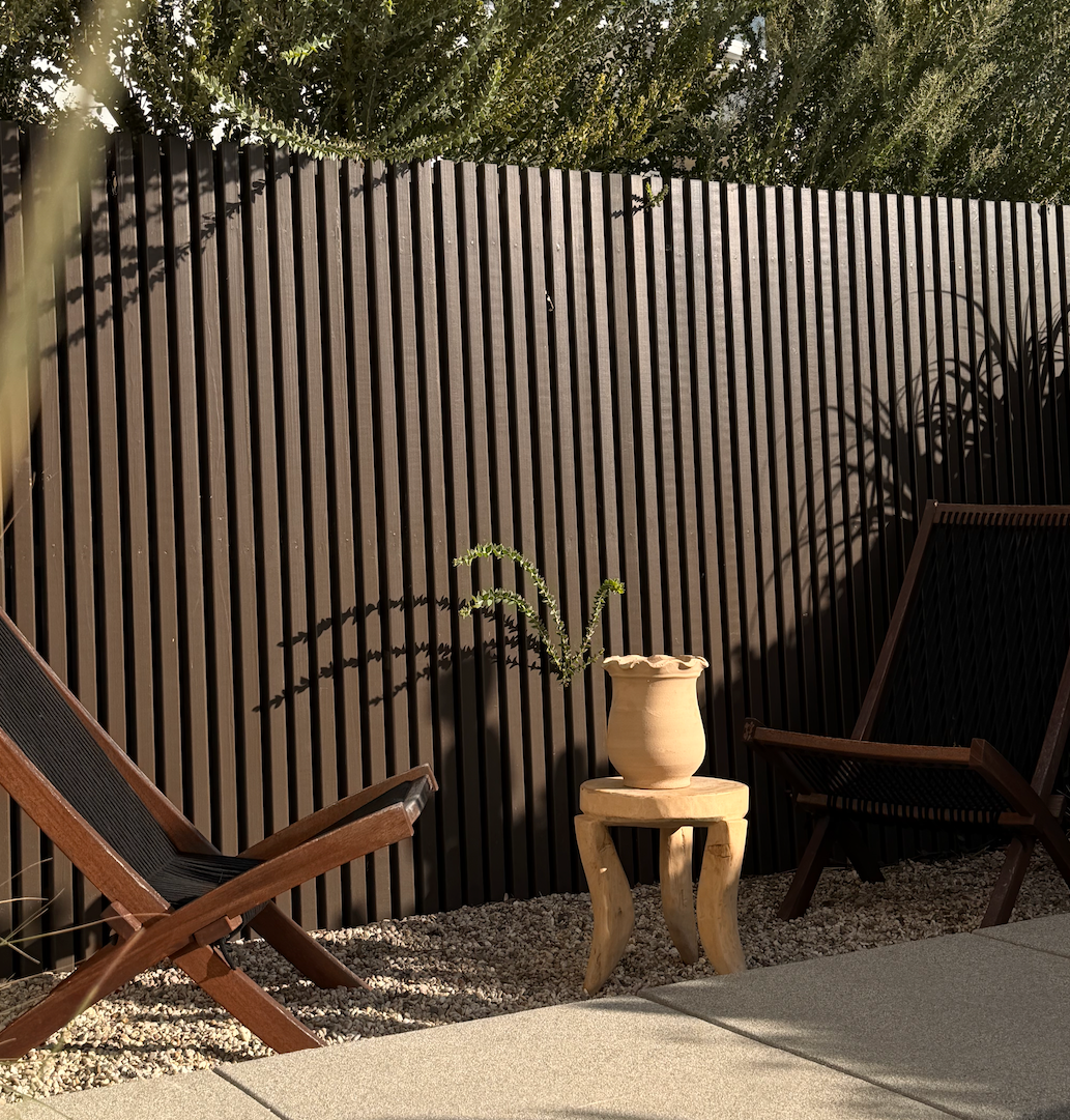 Outdoor setting with wooden lounge chairs and a small table with a plant against a slatted fence.
